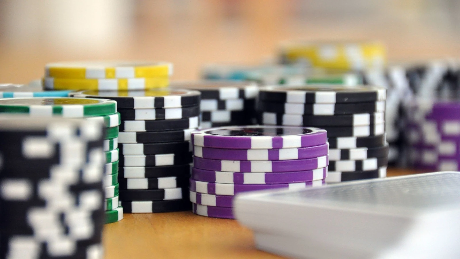 Stacks of tournament poker chips in green, purple, black and yellow denominations on a wooden table, with a deck of cards blurred in the foreground