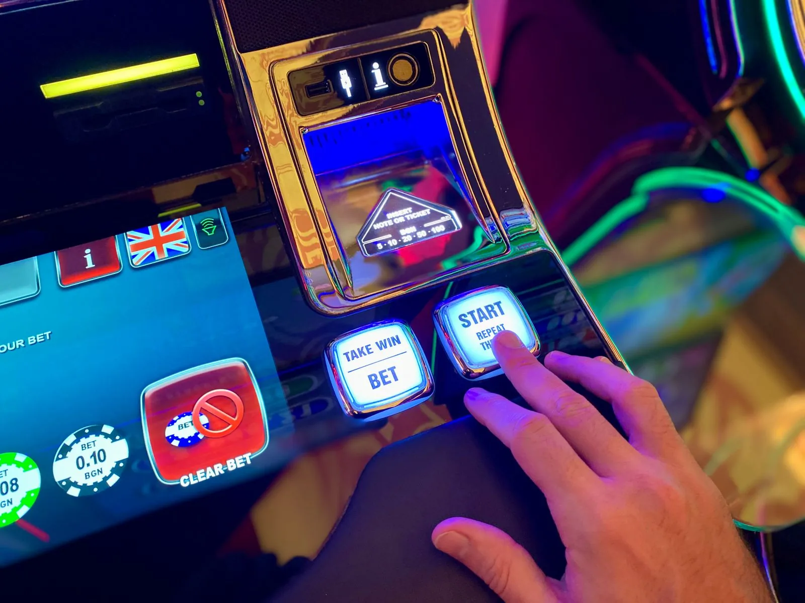 A hand pressing the illuminated start button on a casino slot machine next to a take win and bet button